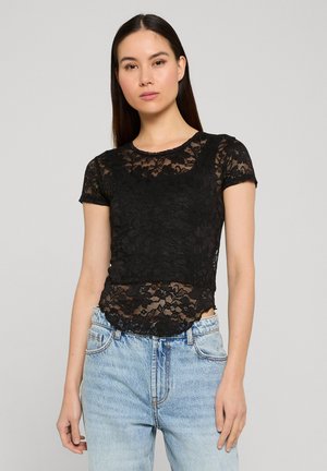 Femme aux longs cheveux bruns portant un haut en dentelle noire à manches courtes avec des motifs floraux et un jean taille haute bleu clair, debout devant un fond uni.