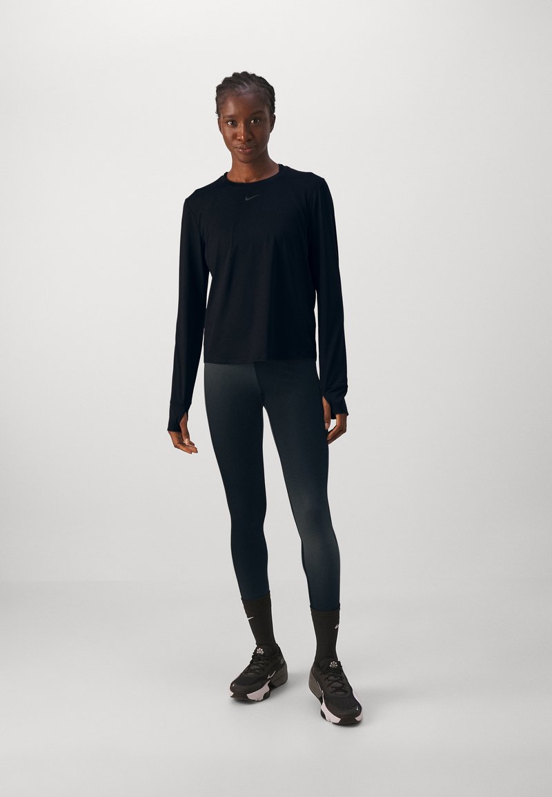 Camiseta atlética de manga larga negra combinada con leggings oscuros y zapatillas de correr negras. Presenta un logo sutil y un diseño ajustado para mayor comodidad.