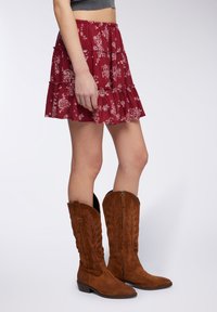 Gonna a balze con stampa floreale rossa e vita elastica, abbinata a stivali da cowboy in camoscio marrone al ginocchio con cuciture decorative.