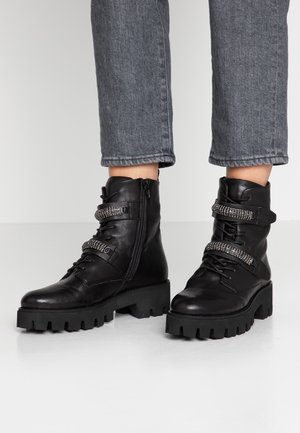 Czarne masywne skórzane botki na grubej podeszwie, z bocznymi zamkami i dwoma ozdobnymi paskami z cyrkoniami, noszone z szarymi krótko skrojonymi dżinsami.