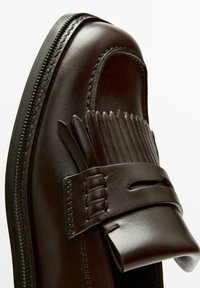 Dark brown leather loafer with fringe detailing and a stitched strap across the upper, shown in close-up against a white background.