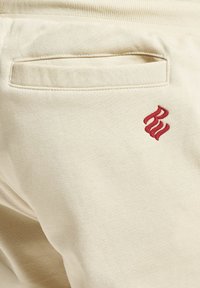 Pantalon beige avec une poche arrière fendue et un petit logo brodé rouge près de la poche sur tissu lisse.
