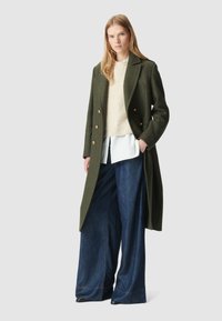 Lungo cappotto verde in lana con bottoni dorati, indossato sopra un maglione lavorato a maglia crema e una camicia bianca, abbinato a pantaloni in denim a gamba larga.