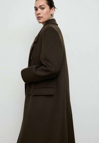 Brązowy, wełniany płaszcz o wyraźnie zarysowanej sylwetce, z dużymi klapami, bocznymi kieszeniami i zapięciem na podwójny guzik. Gładka faktura z minimalnymi zdobieniami.