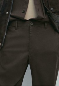 Pantalon marron foncé avec passants pour ceinture, fermeture à boutons, associé à une chemise beige rentrée et une veste marron partiellement visible.