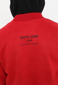 Rode suède jasje met zwarte geribbelde kraag. De achterkant is voorzien van zwarte tekst: "SIXTH JUNE PARIS 48° 51' 57" N, 2° 20' 32" E." Gladde textuur.