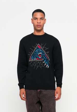 Jonge man in een zwarte trui met een blauw en rood brullende tijgerontwerp die door een grijze driehoekige omlijsting breekt, gecombineerd met vervaagde donkere broek.