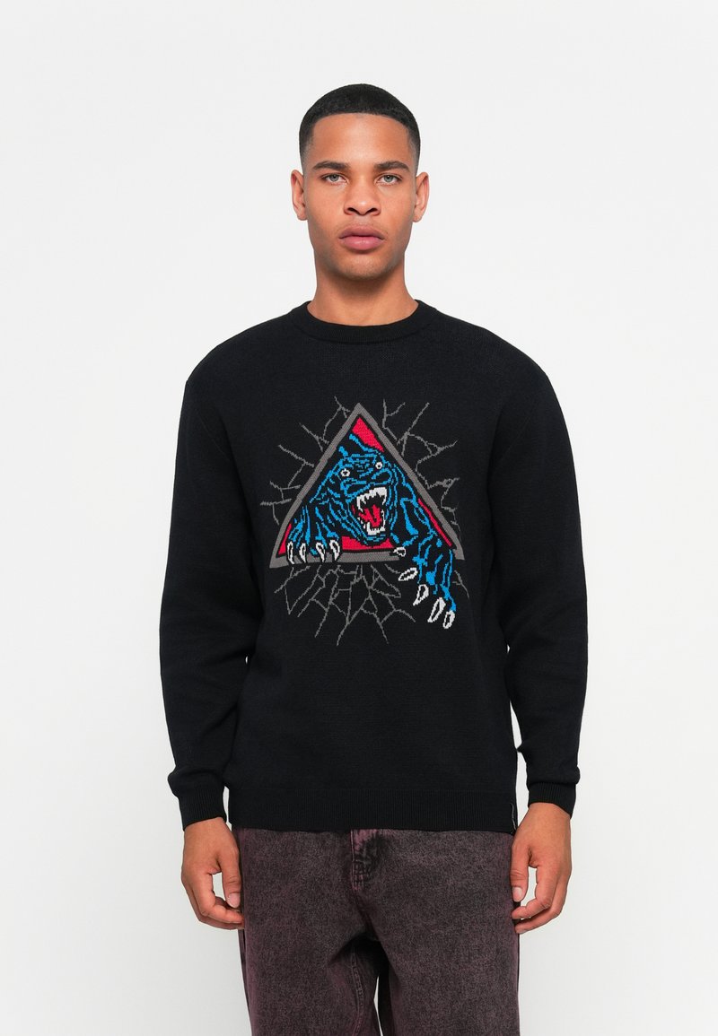 Jonge man in een zwarte trui met een blauw en rood brullende tijgerontwerp die door een grijze driehoekige omlijsting breekt, gecombineerd met vervaagde donkere broek.