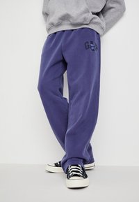 Violeta sporta bikses ar brīvu piegriezumu, kurām ir melns "GAP" logo un gluda tekstūra. Apvienotas ar melniem kanvas kedām.