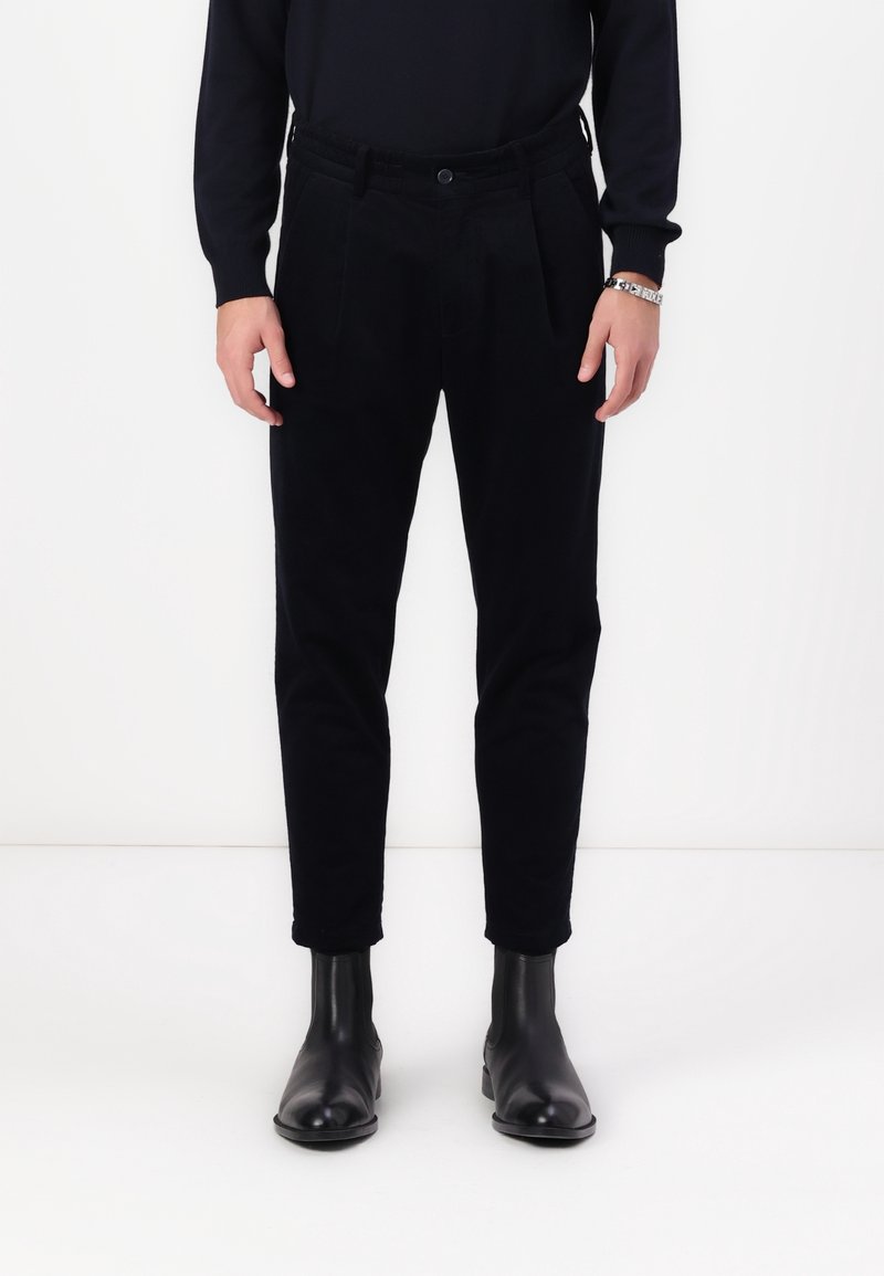 Personne debout sur fond blanc, portant un pantalon noir, un pull noir et des bottes en cuir noires, mains détendues sur les côtés.