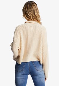 Krämfärgad stickad tröja med lös passform, vida ärmar och kort rygg. Kombinerad med högmidjade blå jeans i denim.