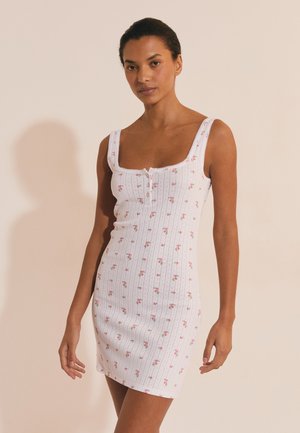 Kobieta o ciemnych włosach spiętych do tyłu, ubrana w białą sukienkę bez rękawów z drobnym różowym kwiatowym wzorem i guzikami, stojąca na jasnym tle.