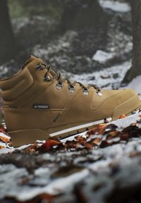 Tan hiking boot with black laces on snow-covered ground with brown leaves and blurred forest background.