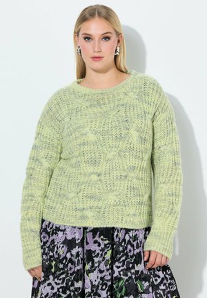 Kobieta ubrana w jasnobrązowy, teksturowany sweter dziany oraz czarną plisowaną spódnicę z fioletowo-zielonymi kwiatowymi wzorami, stojąca na tle gładkiej ściany.