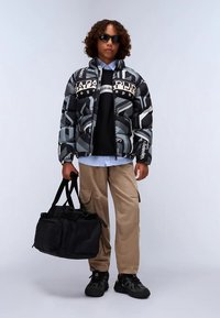 Oseba v vzorčasti črno-sivi pufasti jopici, bež cargo hlačah, črnih teniskah, sončnih očalih, drži črno torbo proti enobarvni ozadju.