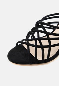 Μαύρο σουέντ σανδάλι με ανοιχτά δάχτυλα και πολλαπλά λεπτά, σταυρωτά λουράκια πάνω σε μπεζ εσωτερική σόλα σε λευκό φόντο.