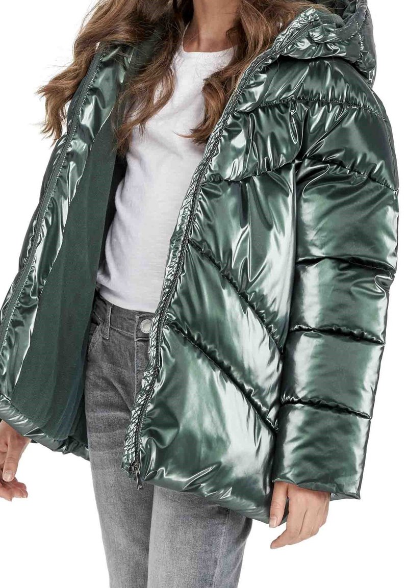 Grøn metallic pufferjakke med hætte, der har lynlåsfront og skrå quiltet mønster. Sammen med en grå top og denim.