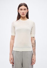 Jasnobeżowy krótki rękaw sweterek z okrągłym dekoltem i prążkowanymi akcentami. Materiał jest przejrzysty, co uwidacznia subtelną teksturę. Noszony z szarymi wzorzystymi spodniami.