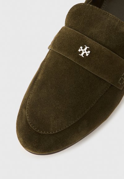 Tumši zaļas zamšādas kurpes bez papēža ar apaļu purngalu, dekoratīvu siksniņu un sudraba logotipa akcentu virspusē. Gluda tekstūra, pieticīgs dizains.