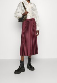 Gonna midi plissettata bordeaux con una texture liscia, abbinata a una chemisier bianca a fantasia e stivali alla caviglia neri con tacco alto e spesso.