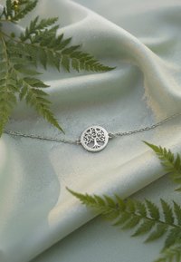 Bracciale in argento a forma di albero della vita con design intagliato intricato, esposto su una morbida stoffa verde chiaro e circondato da felci.