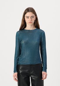 Teal színű, hosszú ujjú felső, texturált, csillogó felülettel és kerek nyakkivágással. Fekete bőr nadrággal párosítva, elegáns megjelenést kölcsönöz.