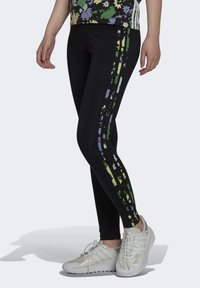 Mustat leggingsit, joissa on vihreät, keltaiset ja purppurat abstraktit sivuraitat, valmistettu venyvästä materiaalista, yhdistetty valkoisiin urheilukenkiin.