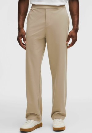 Mann trägt beige, gerade geschnittene Hose und weiße Turnschuhe mit braunen Sohlen, steht vor neutralem Hintergrund.