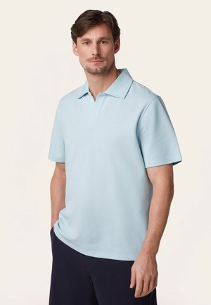 Man draagt een lichtblauw polohemd met korte mouwen en donkere broek, staande met één hand in de zak tegen een effen lichte achtergrond.