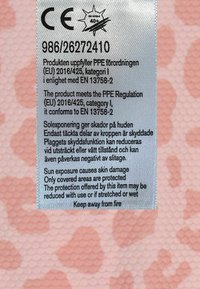 Etikett tryckt på en strukturerad, reflekterande tygbakgrund med rosa mönster, som anger produktens efterlevnad, varningar om sol exponering och skötselråd.