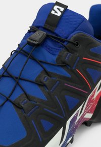 Kék atlétikai cipő texturált szintetikus felsőrésszel, elasztikus fűzőkkel és fekete díszítésekkel. Ívelt talppal rendelkezik, amelyen színátmenetes részlet látható.