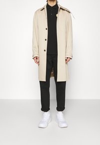 Béžový trenčkot s špičatým límcem, hnědými knoflíky, černé polo tričko, černé kalhoty a bílé tenisky. Minimalistický design, casual styl.