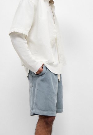 Homme portant une chemise blanche à manches longues superposée avec une chemise à manches courtes, un short en velours côtelé bleu clair, les mains dans les poches.