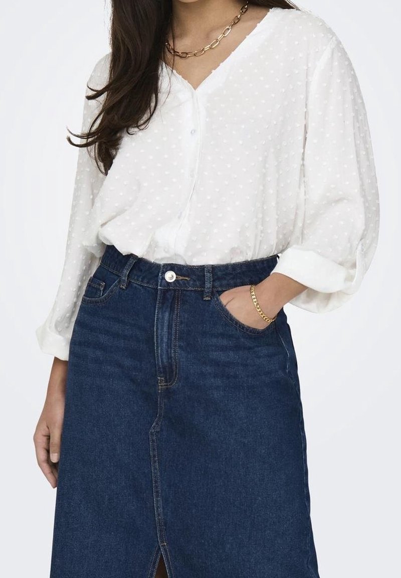 Femme portant un chemisier blanc texturé à manches longues, rentré dans une jupe en jean bleu foncé taille haute avec une fente à l'avant.