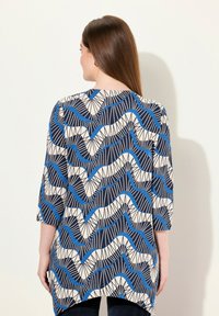 Tunique bleue et crème à motifs, mettant en avant des lignes ondulées et des formes géométriques, avec des manches trois-quarts et une coupe décontractée.