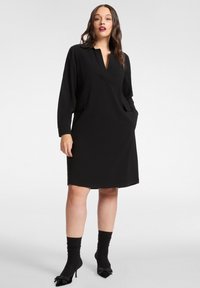 Vestito nero con scollo a V, maniche lunghe e tasche laterali. Tessuto liscio, lunghezza media, abbinato a calzini neri e tacchi. Design semplice.