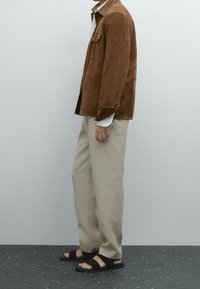 Veste en daim marron, pantalon beige sur mesure, sandales noires. La veste dispose de boutons et de poches, le pantalon a une coupe droite et une texture lisse.