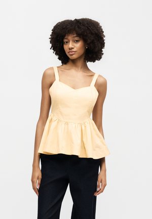 Γυναίκα με κατσαρά μαλλιά που φοράει αμάνικη ανοιχτό κίτρινη μπλούζα τύπου πεπλούμ και μαύρο παντελόνι, στέκεται μπροστά σε απλό λευκό φόντο.