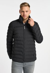 Schwarze Puffjacke mit hohem Kragen, Reißverschluss und vertikaler Steppung, getragen über einem weißen Hemd und kombiniert mit hellen Jeans.
