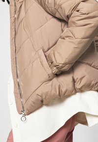 Kurtka puchowa w jasnobrązowym kolorze, z pikowanym wzorem, kieszenią boczną i srebrnym zamkiem. Noszona na białej koszuli z kordurowej tkaniny.