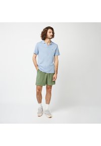 Polo bleu clair à manches courtes, associé à un short vert. Le modèle porte des chaussettes blanches et des baskets claires. Tissu texturé et coupe décontractée.