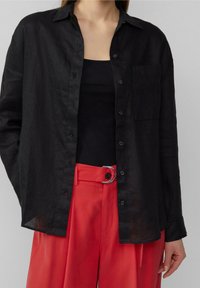 Camisa de lino negra con un diseño de botón, un bolsillo en el pecho y mangas largas, combinada con pantalones rojos de cintura alta que cuentan con una hebilla de cinturón circular.