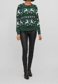 Grön jultröja med vita renar och snöflingor, med texten "GOD JUL" och ribbade fållar, tillsammans med svarta läderbyxor.