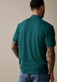 Polo de manga corta en color verde azulado, hecho de una tela suave, con un cuello clásico y un dobladillo recto. El fondo es de un tono neutro.