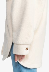 Kremfarget fleecejakke med myk tekstur, oversized silhuett, knappdetalj ved mansjetten, brukt over blå denimjeans.
