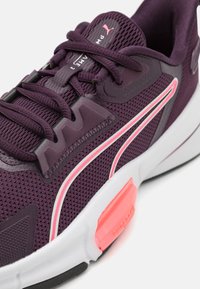 Zapatilla deportiva de malla púrpura con acentos en negro y blanco, ribete rosa y suela de goma texturizada; presenta un cordón prominente y detalles de marca.