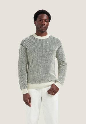 Homme portant un pull en maille à motifs noir et blanc et un pantalon blanc, debout devant un fond clair uni.