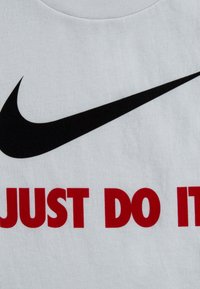 Balta medvilninė marškinė su dideliu juodu Nike swoosh logotipu ir raudonu tekstu, skaitančiu "TIESIOG DARYK TAI" didžiosiomis raidėmis.