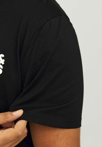 Schwarzes T-Shirt aus Baumwolle mit Rundhalsausschnitt und kurzen Ärmeln. Der Stoff hat eine glatte Textur. Sichtbare Hand, die den Saum hält.