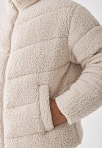 Bež šuplji puffer jakna s kvadratnim dizajnom, sa zatvaranjem na zip, bočnim džepovima i rebrastim manžetama. Mekana teksturirana tkanina.